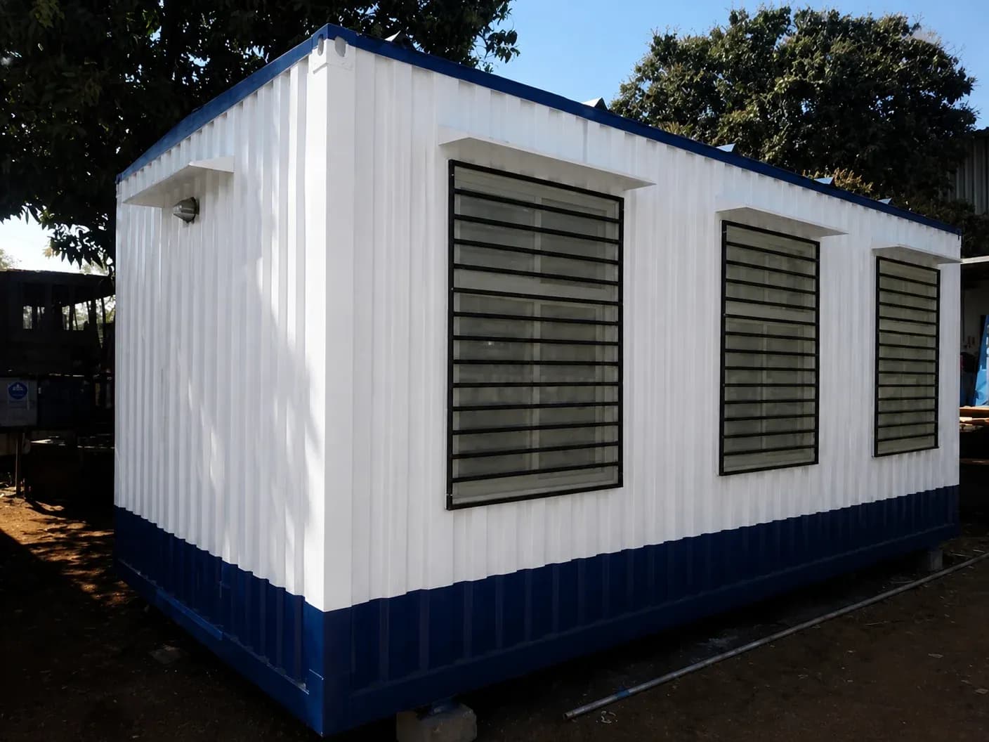 Illustrative example of a white and blue container office cabin seen from one end, with an outdoor wall light and three windows fitted with grilles
