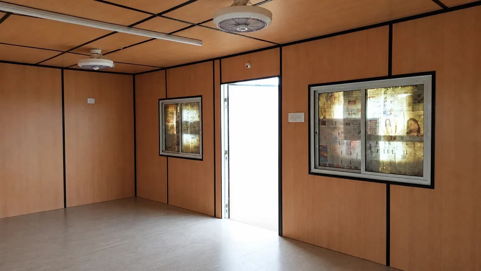 Illustrative example of a container office interior with wood-grain wall panels, two ceiling fans, a tube light and sliding windows