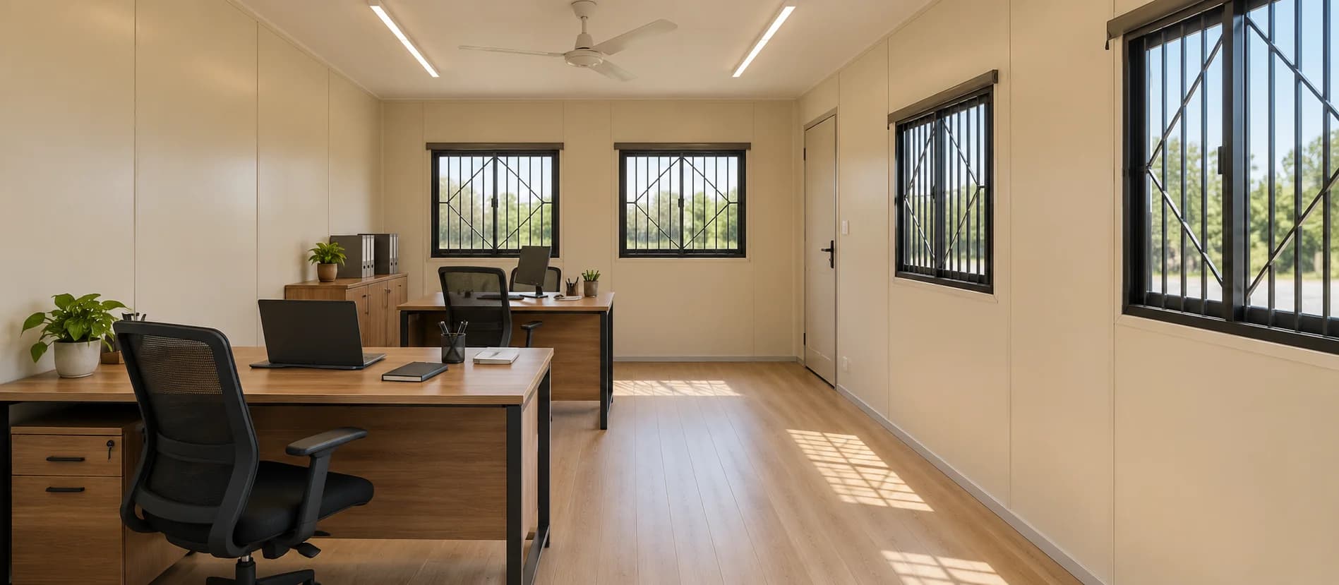 Container office in Sarjapur — optionally furnished interior: cream wall panels, wood-grain laminate flooring, two desks with mesh chairs and a laptop, a storage unit, a ceiling fan, LED tube lights and grilled windows