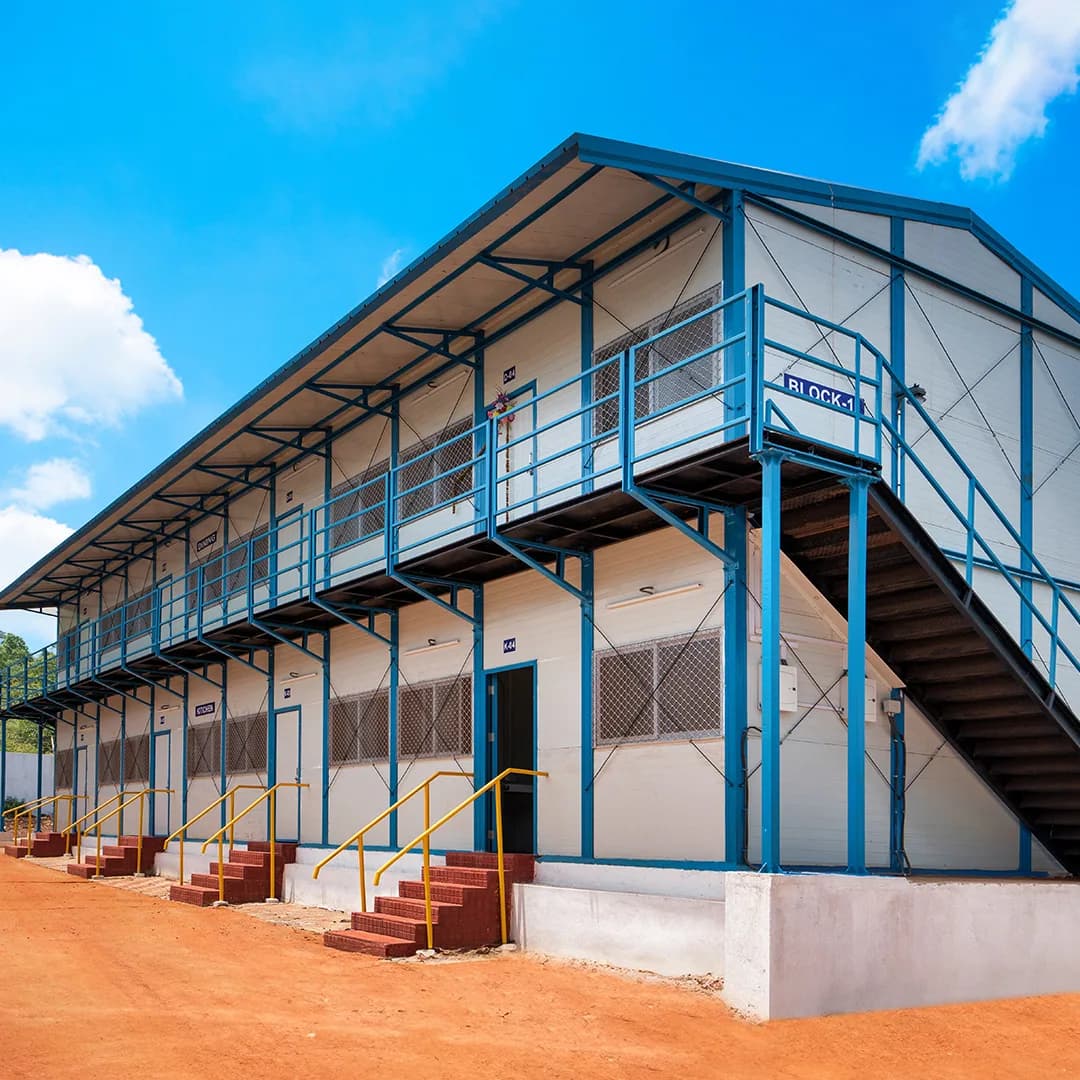 Labour camp setup in Bangalore — G+1 workers' dormitory block with a blue steel frame, external staircase, first-floor walkway, mesh-screened windows and brick entrance steps, signed Block-1