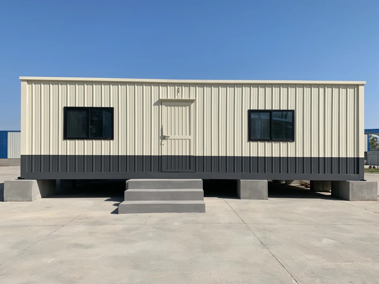 Portable cabin in Kolar — illustrative render of a cream container-style portable cabin with ribbed steel walls, a charcoal lower band, two black-framed sliding windows and a panelled entrance door, standing on concrete blocks with a three-step stair on a paved yard